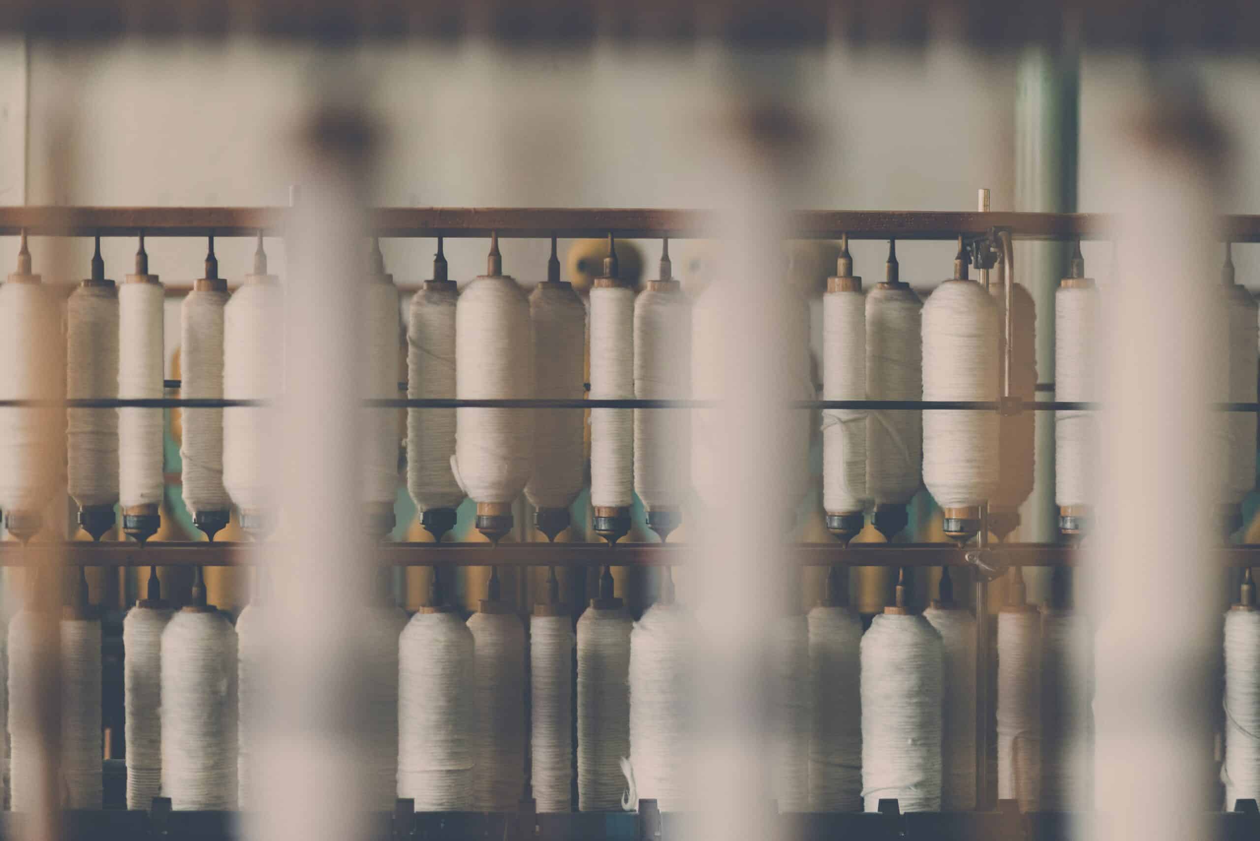 Spools of white thread on an industrial textile machine in a fashion manufacturing facility, illustrating the apparel production roles that fashion recruiters and fashion staffing agencies help fill.