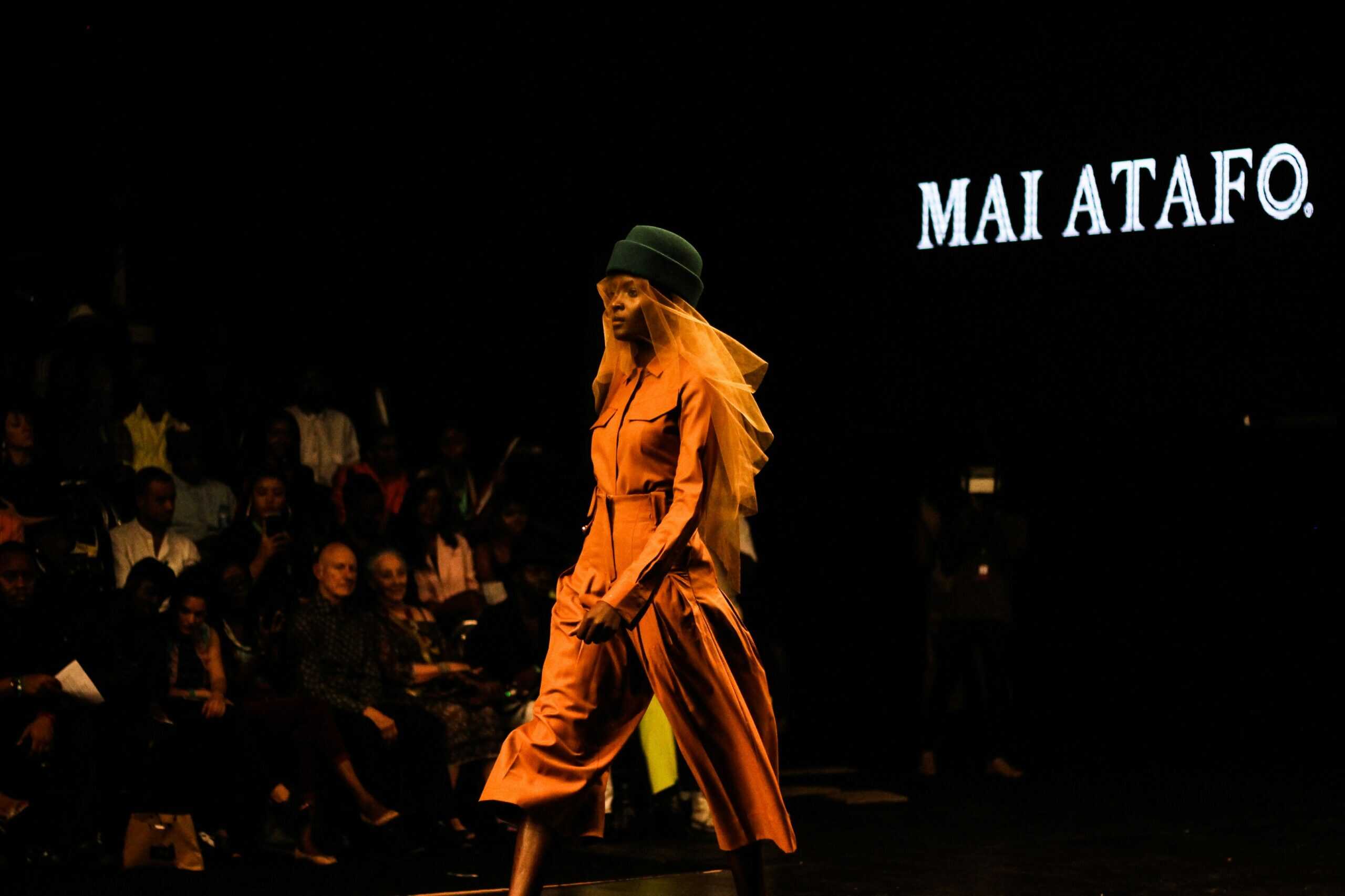 Model walking the runway in an orange jumpsuit and green hat with a yellow veil during a Mai Atafo fashion show, with audience seated in the background.
