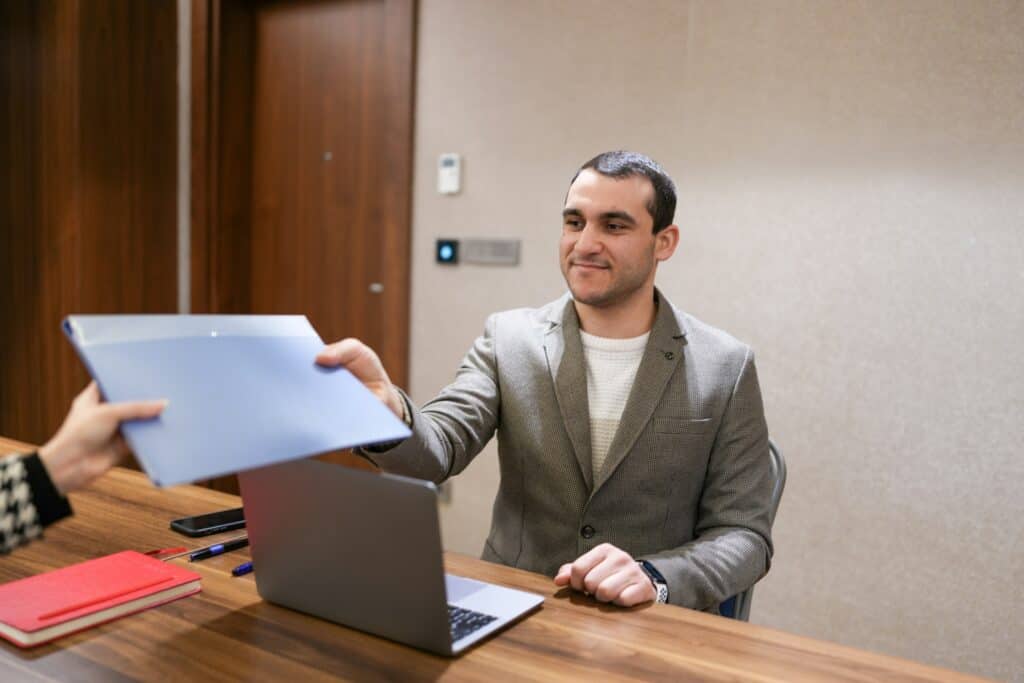 Fashion recruiter in a blazer receives a candidate’s portfolio folder during an interview at an office desk.