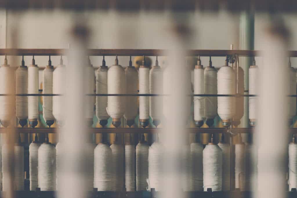 Spools of white thread on an industrial textile machine in a fashion manufacturing facility, illustrating the apparel production roles that fashion recruiters and fashion staffing agencies help fill.